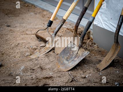 Pale usate sporche e un rastrello appoggiano contro un muro durante una pausa di lavoro in un nuovo cantiere di costruzione casa. Foto Stock
