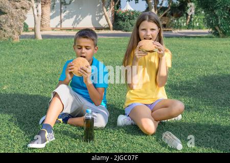 Bambini gli scolari mangiano gli hamburger nel parco seduti sull'erba. Pranzo scolastico. Foto Stock