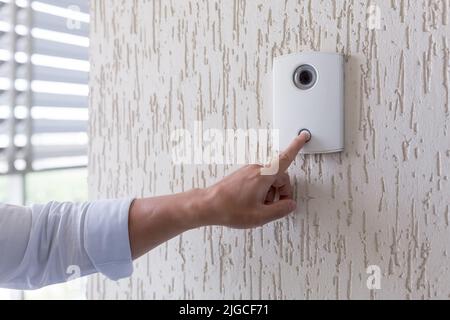 Mano di un uomo in una camicia bianca premendo il pulsante di una porta bussare su una parete bianca di una casa, un intelligente porta bussare con una videocamera per la comunicazione Foto Stock