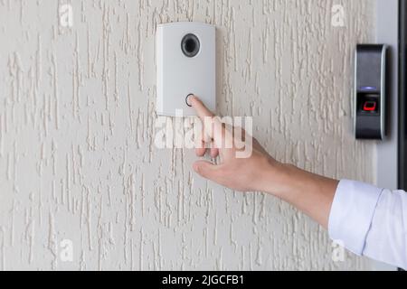 Mano di un uomo in una camicia bianca premendo il pulsante di una porta bussare su una parete bianca di una casa, un intelligente porta bussare con una videocamera per la comunicazione Foto Stock