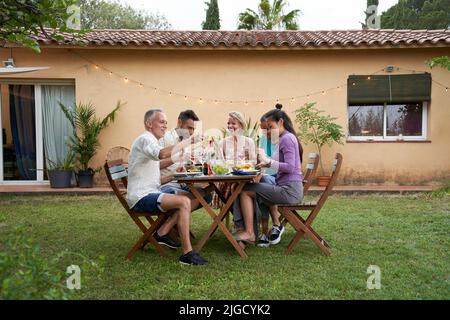 Un gruppo di amici si è riunito intorno ad un tavolo in un giardino nel pomeriggio estivo per condividere un pasto e avere un buon tempo insieme. Foto Stock
