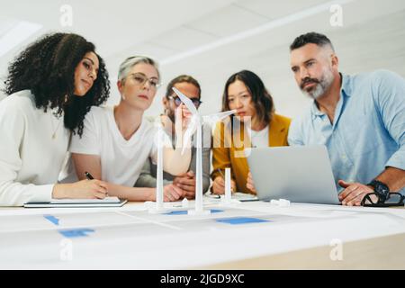 Progettisti multiculturali che lavorano con 3D modelli di turbine eoliche in un ufficio moderno. Team di uomini d'affari innovativi brainstorming mentre si lavora su una ren Foto Stock