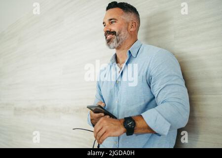 Un uomo d'affari felice e maturo sorridente e che guarda via mentre si trova in un ufficio moderno. Uomo d'affari maturo e premuroso che tiene uno smartphone in piedi Foto Stock
