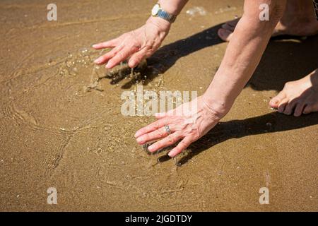 Donna anziana attraente che tocca l'acqua sulla riva del mare Foto Stock