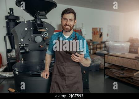 Barista sorridente con telefono in piedi sullo sfondo della macchina per la tostatura del caffè Foto Stock