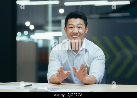 Un uomo d'affari asiatico di successo e felice che guarda la macchina fotografica, parla di videochiamata, gesticolando allegro all'interlocutore, all'incontro online, all'uomo che lavora in ufficio al lavoro Foto Stock