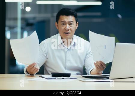 Ritratto di insoddisfatto investitore d'affari, uomo d'affari asiatico che lavora in ufficio, guardando la macchina fotografica e tenendo documenti, deluso con la relazione Foto Stock