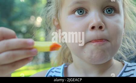 La bambina mangia patatine fritte. Primo piano di ragazza bionda prende patatine con le mani e li prova seduti in strada caffè sul parco. Foto Stock