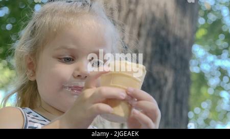 La ragazza piccola carina mangia il gelato fuori. Primo piano ritratto della ragazza bionda seduta sul panchina del parco e mangiare icecream. Foto Stock