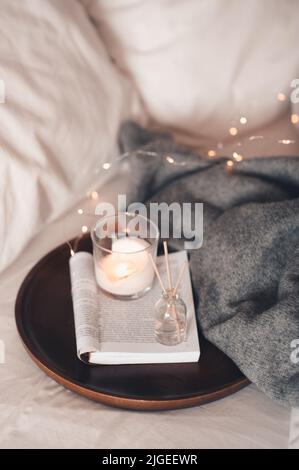 Home fragranza liquida su libro di carta aperto e candela profumata bruciante su vassoio di legno e maglione di stoffa a maglia in letto da vicino. Atmosfera accogliente. Foto Stock