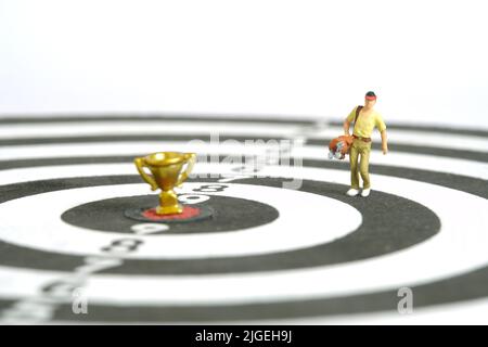 Fotografia di personaggi giocattolo in miniatura. Un caddy da golf in piedi sopra la tavola da disegno con trofeo d'oro al centro, isolato su sfondo bianco. Immagine pho Foto Stock