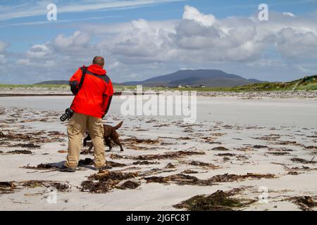 Un uomo di mezza età in una giacca rossa brillante che gioca con un cane su una spiaggia di sabbia bianca a South Uist Foto Stock