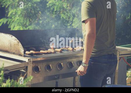 Guy cucina salsicce sul barbecue grill, il fuoco aperto in una giornata estiva nel cortile di una casa privata. Foto Stock