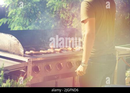 Guy cucina salsicce sul barbecue grill, il fuoco aperto in una giornata estiva nel cortile di una casa privata. Foto Stock