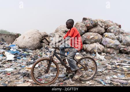 Un povero ragazzo africano di slum passa in bicicletta attraverso la discarica della città in cerca di vecchi pneumatici di scorta Foto Stock