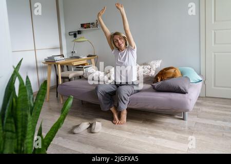 Donna bionda di mezza età ha iniziato la sua mattina a letto con il cane e stretching con piacere sorriso Foto Stock