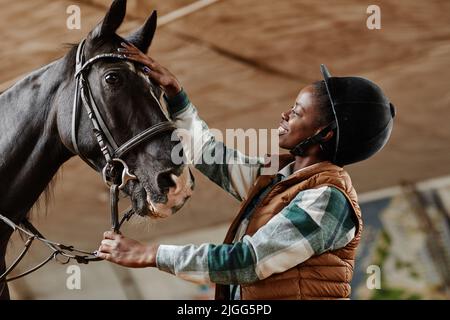 Vista laterale ritratto di sorridente donna afroamericana che stroking cavallo nero in campo di equitazione al coperto in pratica Foto Stock