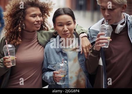 Ragazzo giovane che beve soda attraverso sraw mentre cammina con due ragazze interculturali felici o torna a casa dopo l'università Foto Stock