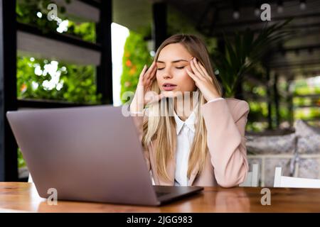 Donna stressata usando il laptop e ha mal di testa fuori in un bar. Ragazza seduta sul tavolo e toccare la testa, ha forte mal di testa, mentre il lavoro o Foto Stock