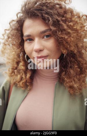 Giovane bella donna con capelli lunghi e spessi marroni ondulati e trucco naturale guardando la macchina fotografica mentre si trova in piedi per strada Foto Stock