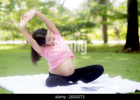 Felicità giovane donna asiatica incinta seduta facendo esercizio sportivo con mani stretch muscolo per la salute al parco, madre che pratica yoga per il leisur Foto Stock