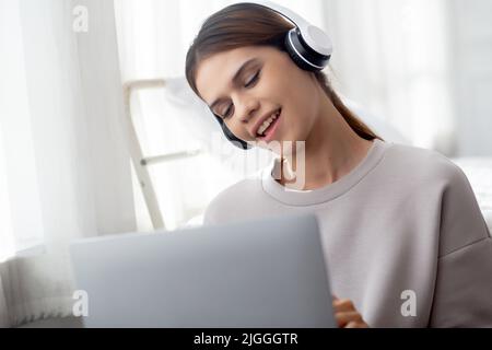 Felice giovane donna asiatica che indossa le cuffie ascoltare musica mentre si utilizza il computer portatile e cantare con divertimento e godere nel soggiorno, felicità fe Foto Stock