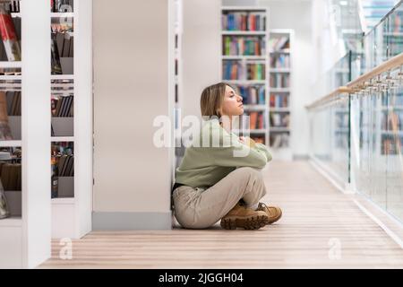 Donna pensiva pensiva di mezza età con libro in mani seduto sul pavimento in una moderna biblioteca interna Foto Stock