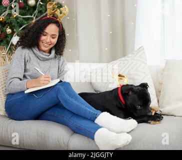 Fare una lista di alcuni acquisti di Natale dell'ultimo minuto. Una donna che scrive nel suo taccuino mentre seduta a casa con il suo cane durante il periodo di Natale. Foto Stock