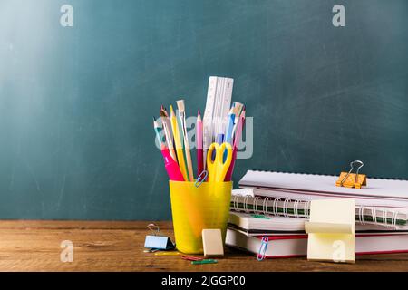 Molte diverse scuole, forniture per ufficio e cancelleria. Concetto di ritorno a scuola. Taccuino, libro, penna, matita, forbici, sveglia, orologio, stativo adesivo su un tavolo di legno su sfondo verde. Foto Stock