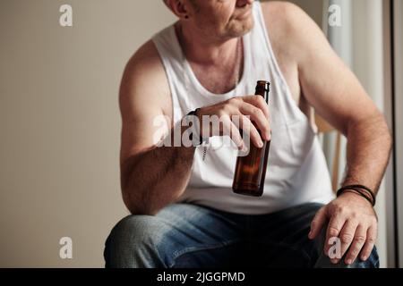 Immagine ritagliata dell'uomo maturo in camicia bianca che beve birra e guarda all'esterno attraverso la finestra Foto Stock