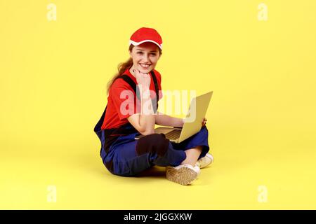 Ritratto completo di donna da lavoro ottimista seduta sul pavimento con gambe incrociate e lavoro sul notebook, guardando la fotocamera e tenendo il mento. Studio interno girato isolato su sfondo giallo. Foto Stock