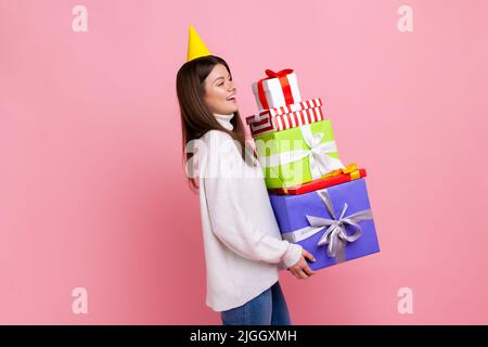 Ritratto laterale di donna felice in cono festa con un sacco di scatole regalo in mani, festeggiamenti compleanno, indossando bianco stile casual maglione. Studio interno girato isolato su sfondo rosa. Foto Stock