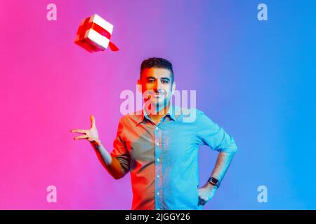 Ritratto di uomo in camicia in piedi con felice espressione facciale positiva e mano sollevata, cercando di catturare il regalo volante. Studio interno girato isolato su colorato sfondo di luce al neon. Foto Stock