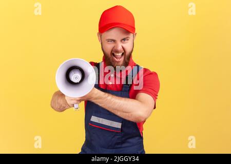 Ritratto di uomo operaio pazzo eccitato che indossa la uniforme blu e il cappuccio rosso, tenendo il megafono e guardando la fotocamera con volto accigliato. Studio interno girato isolato su sfondo giallo. Foto Stock