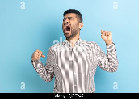 Ritratto di giovane uomo d'affari attraente con barba che sbadigia e solleva le mani, si sente stanco di lavoro straordinario, indossando una camicia a righe. Studio interno girato isolato su sfondo blu. Foto Stock