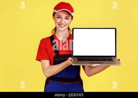 Ritratto di donna lavoratrice sorridente positiva in piedi e mostrare un laptop con display vuoto con spazio per il testo, indossare tute e cappuccio rosso. Studio interno girato isolato su sfondo giallo. Foto Stock