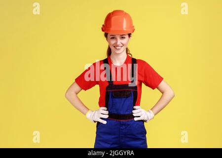 Ritratto di donna costruttore sorridente e sicura in piedi tenendo le mani sui fianchi, guardando la macchina fotografica, indossando tute, guanti e casco protettivo. Studio interno girato isolato su sfondo giallo. Foto Stock