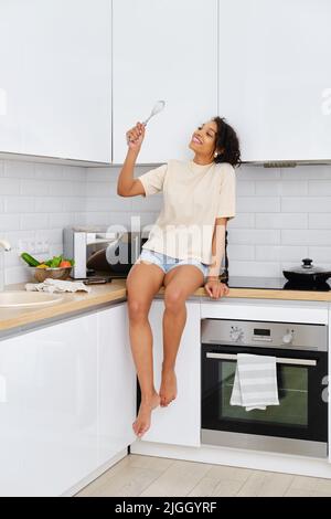 Giovane donna afroamericana sorridente si siede sul tavolo nella cucina sussurra per battere le uova Foto Stock