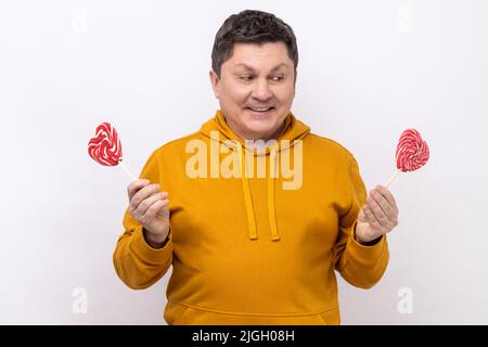 Ritratto di uomo allegro che tiene dolce cuore a forma di caramella lecca e guardando con felice espressione positiva, indossando felpa con cappuccio urban style. Studio interno girato isolato su sfondo bianco. Foto Stock