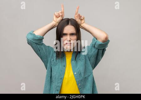 Ritratto di donna aggressiva bully con capelli scuri che mostrano corna toro gesto sopra la testa, accigliando come prima attacco, indossando casual giacca stile. Studio interno girato isolato su sfondo grigio. Foto Stock