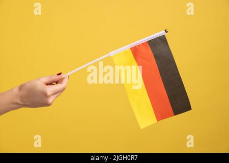 Profilo vista laterale primo piano della mano donna che tiene bandiera della Germania. Giorno della Germania - 3th ottobre. Studio interno girato isolato su sfondo giallo. Foto Stock