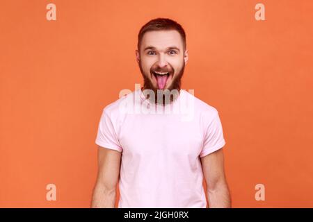 Ritratto dell'uomo barbuto che mostra la lingua e guarda la macchina fotografica con gimace disobbediente, facendo il viso, indossando una T-shirt rosa. Studio interno girato isolato su sfondo arancione. Foto Stock