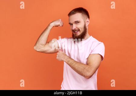 Ritratto di orgoglioso uomo con bearded forte che punta a mano rialzata che dimostra bicipite, sicuro nel corpo sensazione potente, indossando T-shirt rosa. Studio interno girato isolato su sfondo arancione. Foto Stock