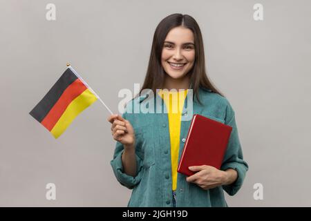 Ritratto di attraente donna ottimista intelligente che tiene in mano libro e bandiera tedesca, corsi di istruzione all'estero, indossando giacca casual stile. Studio interno girato isolato su sfondo grigio. Foto Stock