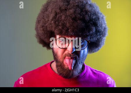 Ritratto di un uomo hipster con acconciatura Afro che mostra la lingua con grimace disobbediente naughty, facendo il viso, indossando una felpa rossa. Studio interno girato isolato su colorato sfondo di luce al neon. Foto Stock