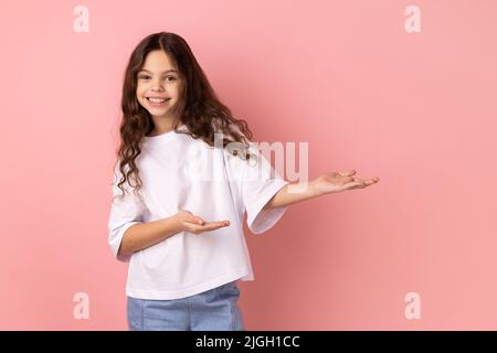 Ritratto di generosa ragazza generosa gentile-hearted che indossa la T-shirt bianca che mostra il gesto di benvenuto e lo spazio vuoto sulla parete per la vostra pubblicità migliore. Studio interno girato isolato su sfondo rosa. Foto Stock