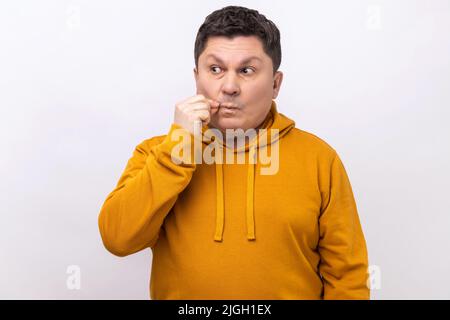 Non dirò a nessuno, prometto. Uomo di mezza età che fa un gesto di zip per chiudere la bocca, mantenere segreto, zip labbra, indossando una felpa con cappuccio urban style. Studio interno girato isolato su sfondo bianco. Foto Stock