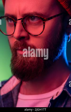 Ritratto closeup di stretto maschio bossy bearded in occhiali che guardano allo spazio laterale con faccia seria attenta, indossando cappello di beanie. Studio interno girato isolato su colorato sfondo di luce al neon. Foto Stock