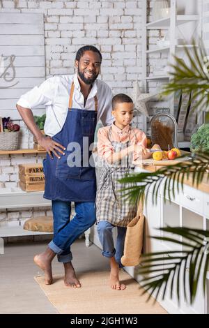 Il padre nero e il bambino che cucinano verdure fresche in cucina a casa. Papà africano e bambino ragazzo che si preparano insieme. Foto Stock