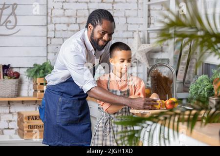 Il padre nero e il bambino che cucinano verdure fresche in cucina a casa. Papà africano e bambino ragazzo che si preparano insieme. Foto Stock